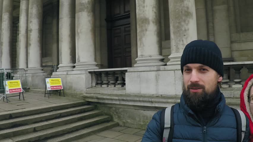 A guy in a dark blue hat and jacket with a beard and long hair walks in a park in London near a colonnaded building with a girl in a red down jacket with a capishon and smiling