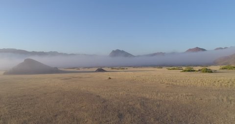 4K tilt up aerial view over dry grasslands and into a dry river bed with fog around mountains,Hoanib Valley, Namib Desert, Namibia