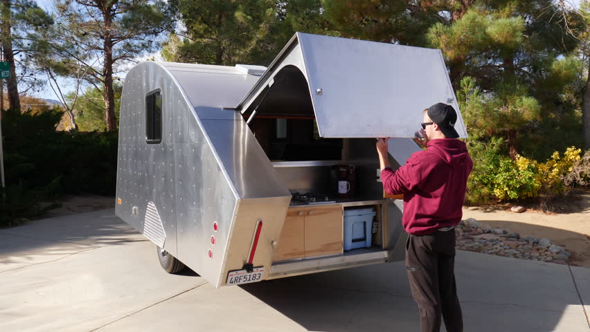 A man opens a deluxe metal teardrop travel trailer in slow motion to get to the stove and kitchen area while camping in his forest campsite.