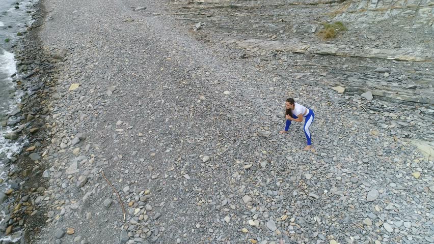 Girl is doing exercise for legs the bodyflex during breathing exercises on the sea and rock background. Aerial video.