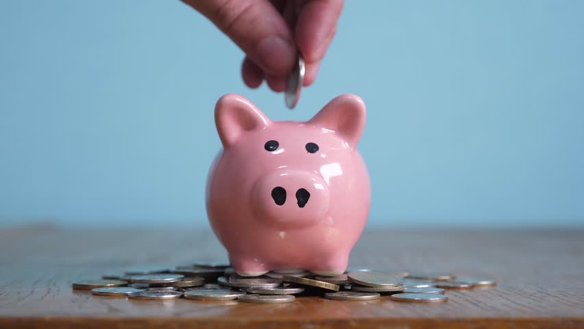 Piggy bank business standing on a pile of coins concept. A hand is putting coin in a piggy bank on a yellow background. saving money is an investment for the future. Banking investment and finance | Shutterstock HD Video #1024048943