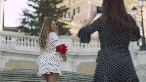 Two young tourists taking pictures with a phone on the Spanish Steps with a bouquet of red flowers with soft natural light. Wide shot on 4k RED camera.
