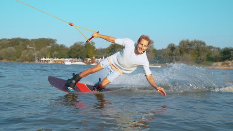 SLOW MOTION, CLOSE UP: Cheerful young man smiles at the camera as her wakesurfs at the cable park on the calm lake. Happy Caucasian guy riding his wakeboard and speeding on the tranquil water surface.