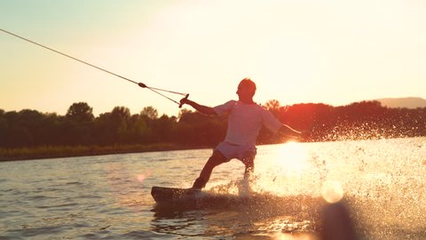 SLOW MOTION, CLOSE UP, LENS FLARE: Happy man having fun wakeboarding in the cool cable park at sunrise. Cinematic shot of a cheerful young guy splashing water while wakesurfing on the tranquil lake.