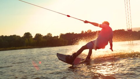 SLOW MOTION, CLOSE UP, SUN FLARE: Unrecognizable athletic man wakesurfing on the lake at golden sunset. Young surfer dude having fun wakeboarding in the cable park on a beautiful summer morning.