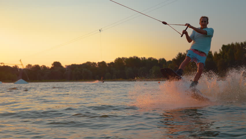SLOW MOTION, TIME REMAP, SUN FLARE, CLOSE UP: Cool surfer dude does 180 ollie while wakeboarding on a sunny summer evening. Cinematic shot of a cheerful man wakeboarding in the cool cable wake park Royalty-Free Stock Footage #1024051934