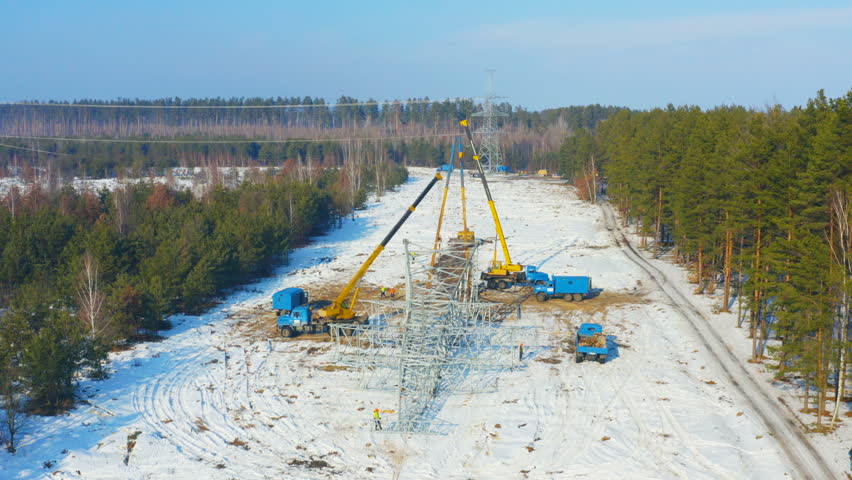 Aerial view of erecting of the power transmission lines, power line pylon (transmission or power tower) by steel erector workers in the forest on a winter day