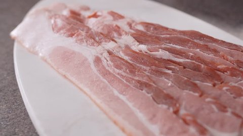 Fresh raw sliced bacon pieces on white cutting board in the kitchen. Camera moving slowly over a closeup view of raw, uncooked bacon slices on the cutting board.