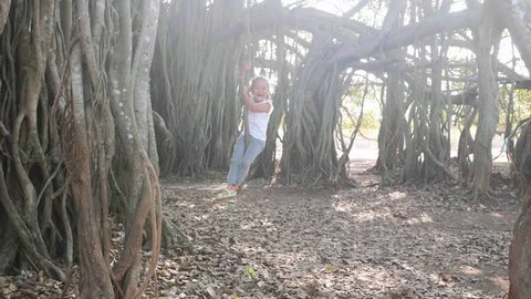 Little cute child girl is laughing and swinging on liana under banyan tree in slow motion. Kid having fun outdoor.
