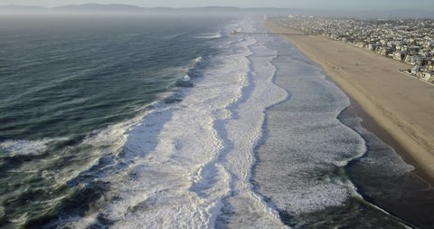 Aerial wide shot POV of California coastline and whitewater waves crashing on the beach from a helicopter view showing the ocean community and pier in Manhattan Beach, Los Angeles, United States