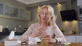 Medium shot of blond woman with sore throat sitting at kitchen table and coughing while talking to doctor on video call