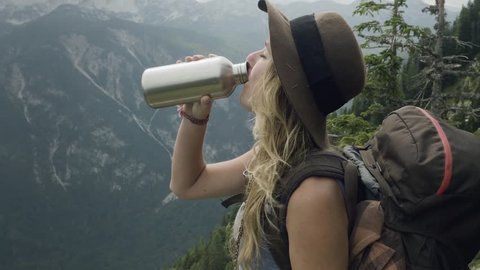 Blonde girl in the mountains with hat drinking water from stainless steel bottle while enjoying the mountain view of slovenian alps