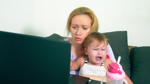 Annoyed businesswoman holding a crying baby in her arms while working on a laptop. Career mom concept, problems associated with work at home