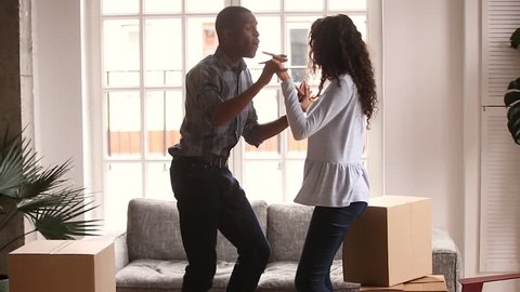 Happy african american first time home buyers couple dancing laughing having fun celebrate relocation buy house in living room with boxes, cheerful black family enjoy moving to music in new apartment