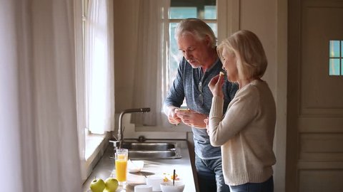 Happy loving senior mature couple having fun preparing healthy breakfast food in kitchen, playful old aged smiling family talking laughing cooking cutting feeding each other together at home