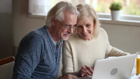 Happy old middle aged couple using laptop talking together doing online shopping, senior mature retired family reading discussing internet computer news, choosing travel offer on website at home