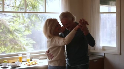 Happy mature senior couple dancing laughing in the kitchen, beautiful romantic middle aged older grandparents relaxing having fun together at home celebrating anniversary enjoy care love tenderness