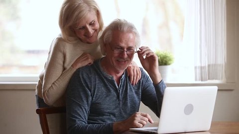 Happy senior middle aged couple laughing talking embracing using laptop together looking at computer screen, smiling older mature family enjoying watching funny video or making online call at home
