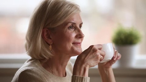 Calm happy mature old woman sitting alone holding cup enjoying aroma morning hot coffee and dreaming, beautiful dreamy senior middle aged lady drinking tea relaxing at home on pleasant leisure day