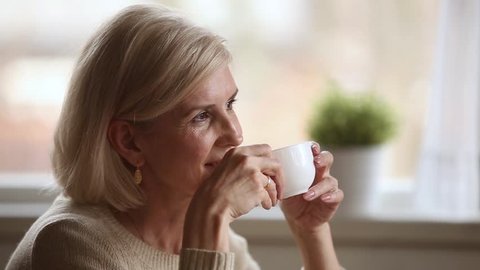 Dreamy happy middle aged mature woman looking away drinking morning coffee or tea at home, smiling senior old lady holding cup relaxing with positive thoughts dreaming enjoying pleasant memories