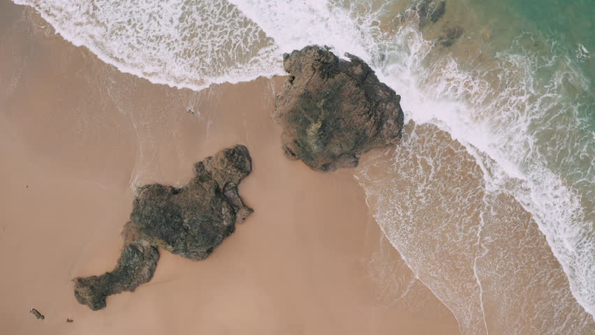 Aerial top down view of wide beautiful beach with big waves and surfers;drone flight over the sandy beach; giant rocks in the middle of the beach; camera moves away; Royalty-Free Stock Footage #1024109546