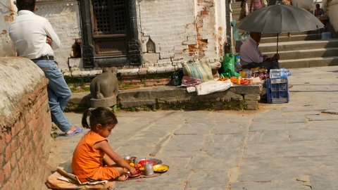 KATHMANDU, NEPAL - 8 OCTOBER 2018 Side view of Little ethnic girl selling various religious attributes, sitting on stone pavement of temple. ghats in Pashupatinath. hindu traditional custom