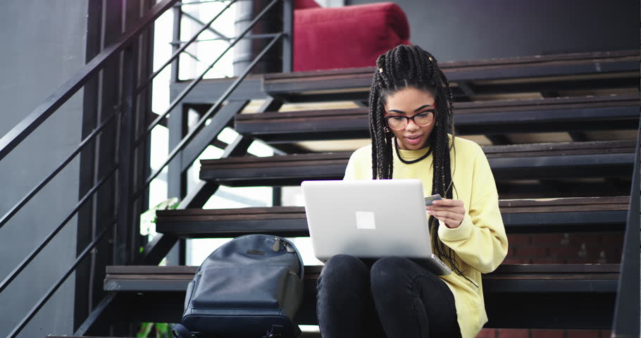 Office worker young african lady with long dreadlocks using her notebook to check the internet banking or to buy something online from her bank card , she sitting down to the stairs in a modern office
