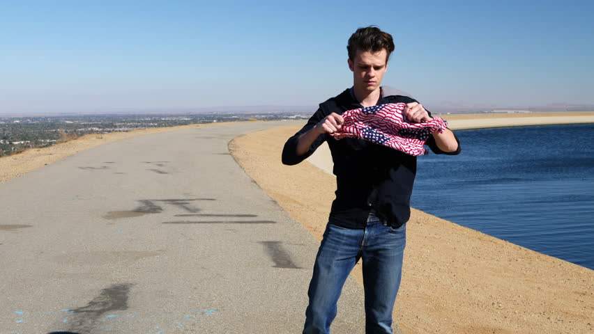 An attractive young man putting on a thief or bandit mask from an american flag bandana in slow motion next to a river in the desert.