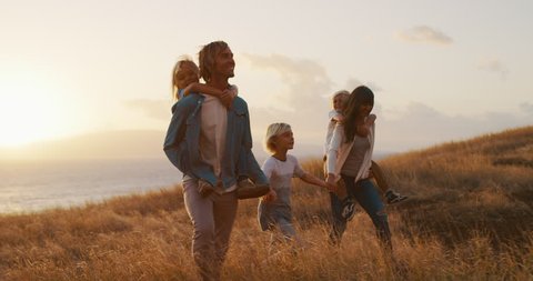 Happy smiling family holding hands walking through golden field at sunset by the ocean, piggy back ride Stock-video