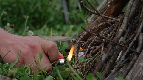 SLOW MOTION, MACRO, DOF: Man's hand holds a lighter and ignites a campfire set up in the wilderness. Unrecognizable man lights up a campfire during a camping trip. Traveler igniting paper and kindling