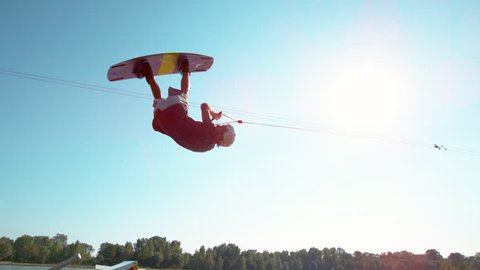 SLOW MOTION, LENS FLARE: Athletic young male wakeboarder does a backflip across the blue sky. Spectacular slow motion shot of extreme athlete doing a cool trick while wakesurfing in a water park.