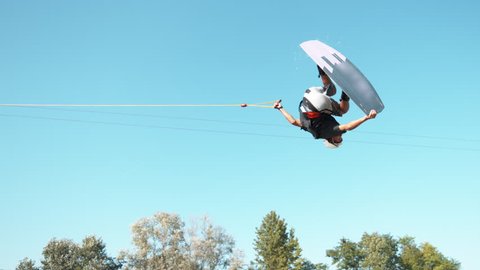 SLOW MOTION: Cool wakesurfer does a backflip and lands on the tranquil lake while being pulled around the water park by a cable. Young male athlete doing cool tricks while wakeboarding in water park.
