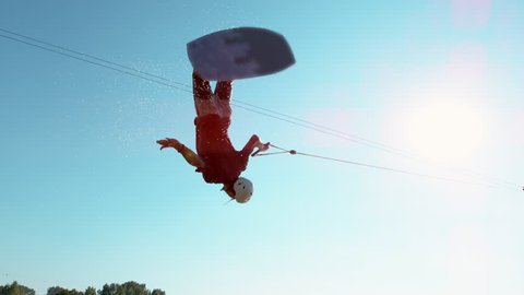 SLOW MOTION, SUN FLARE: Cinematic shot of an athletic young man flipping over the sun while wakeboarding in the fun cable park on the tranquil lake. Extreme athlete having fun wakeboarding in summer.