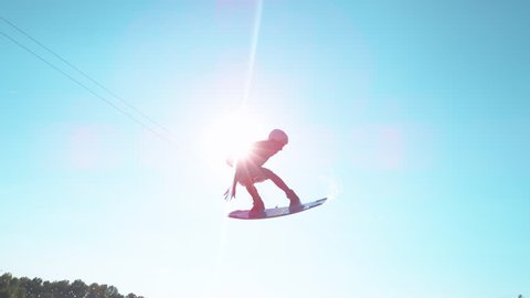 TIME WARP, LENS FLARE: Athletic young man spinning in the air while wakesurfing on the lake. Cinematic shot of bright summer sunbeams shining on extreme wakeboarder doing tricks in the water park.