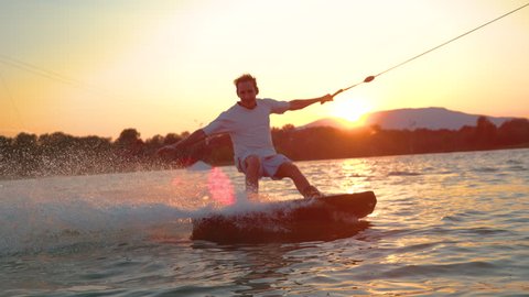 SLOW MOTION, CLOSE UP, LENS FLARE: Cheerful male wakeboarder splashes the refreshing lake water at the camera at sunset. Cinematic shot of a fit young man windsurfing during his active summer vacation