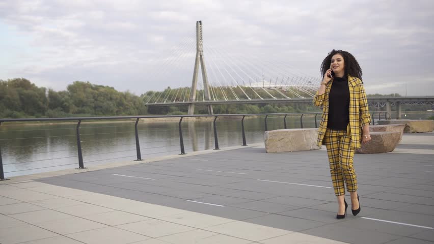 Businesswoman with curly hair walking and talking over the phone next to vistula river and swietokrzyski bridge in the background (gimbal moving shot)