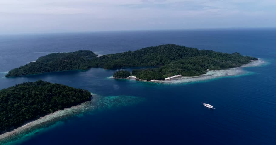 Drone footage of Raja Ampat, Indonesia with a liveaboard boat anchored near Janggelo or Yanggelo island, turquoise water and shallow coral reef around. 