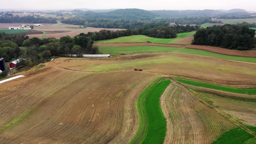 Aerial flyover fallow fields an Amaih man stands on a horse-drawn wagon to guide team view high angle Concept: fall, autumn, traditional, rural, Pennsylvania, countryside, Amish