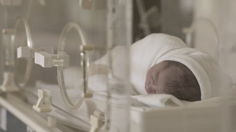 Premature newborn baby with black hair sleeping during recovery in incubator after childbirth. Child moving body, view through the blurred glass, 