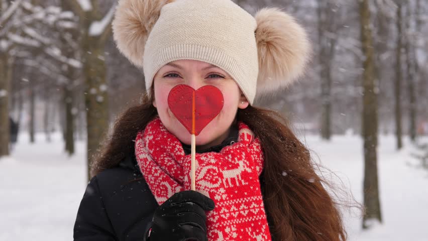 Cheerful woman kiss heart shaped candy and stretch it towards camera. Happy girl portrait at winter outdoors, blurred background. Snow flakes fly around, beautiful weather