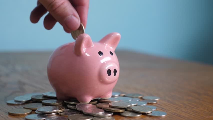 Piggy bank business standing on a pile of coins concept. A hand is putting a coin in a piggy bank on lifestyle a yellow background. saving money is an investment for the future. Banking investment | Shutterstock HD Video #1024169963