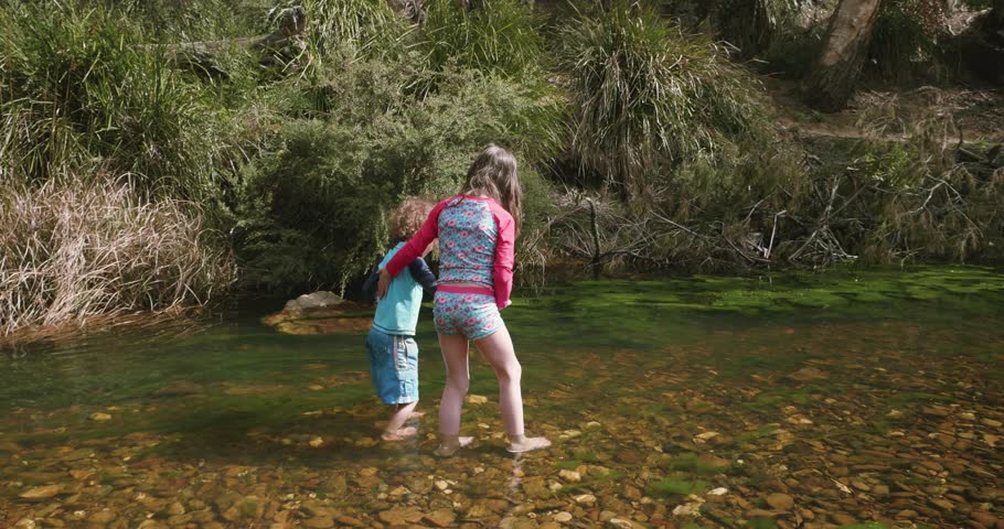 Young girl supports her young brother as they walk knee deep in water in a river with clear water lots of stones and green algae