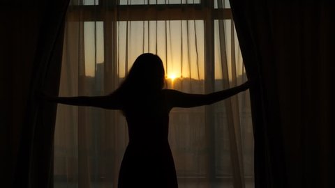 Woman open dark curtains and look to neat sunrise at early hour, black half length silhouette of slender lady against window. She stretch out hands to unveil hangings, slowly and unhurried mood at day