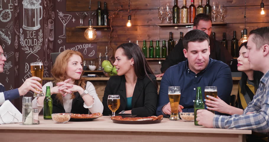 Group of friends clinking their beers in stylish pub or restaurant. They eat pizza and drink beer. A bearded bartender is working in the background.
