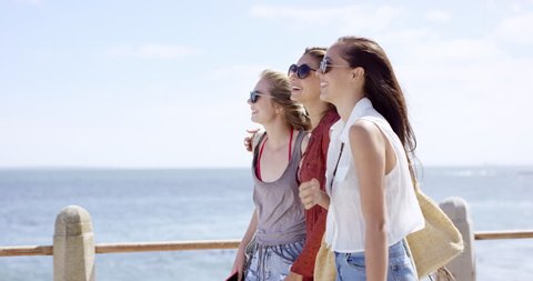 Three young women tourists on summer vacation walking on beach promenade wearing denim shorts