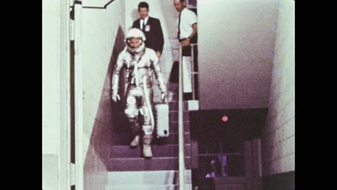 1960s: Astronaut puts a helmet over his head and adjusts it. People walk down the stairs behind astronaut who carries a suitcase, he smiles and waves. Vehicles drive at night, building with NASA logo.