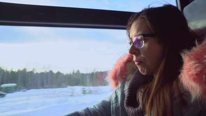 Pensive dark-haired young woman with glasses drinks water and looks out the window during a winter journey by train.