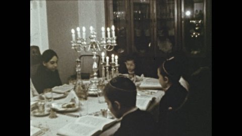1970s: Family at dining table read from Torah. Man stands with chalice of wine near boy with candle. Family gathers around man. Man smells chalice and passes it to children. Children smell chalice.