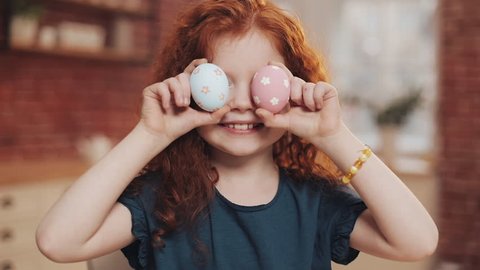 Portrait of cheerful redhead little kid girl playing with easter egg on the kitchen background. She is cheering and having fun at the camera. Happy Easter