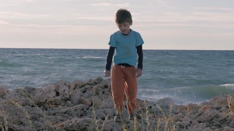 child orphan in dirty clothes on the stony shore. little refugee boy playing on the run of the sea. concept migration and lifestyle